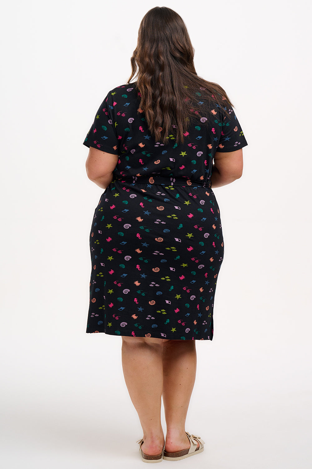 A model wearing a short-sleeved black mini dress with a V-neck, featuring a multicolored underwater theme print. The dress has a matching tie belt and pockets on the skirt.