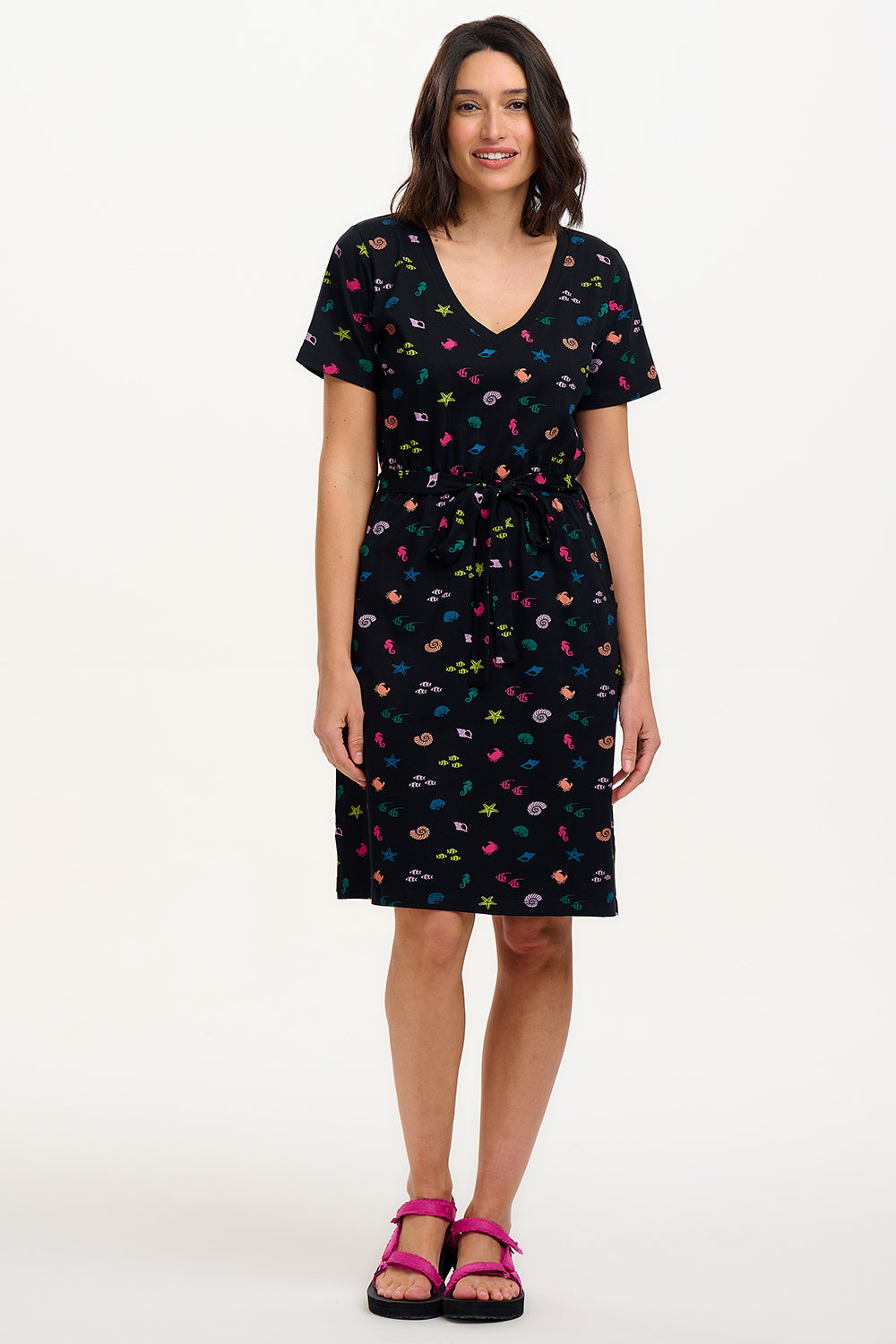 A model wearing a short-sleeved black mini dress with a V-neck, featuring a multicolored underwater theme print. The dress has a matching tie belt and pockets on the skirt.