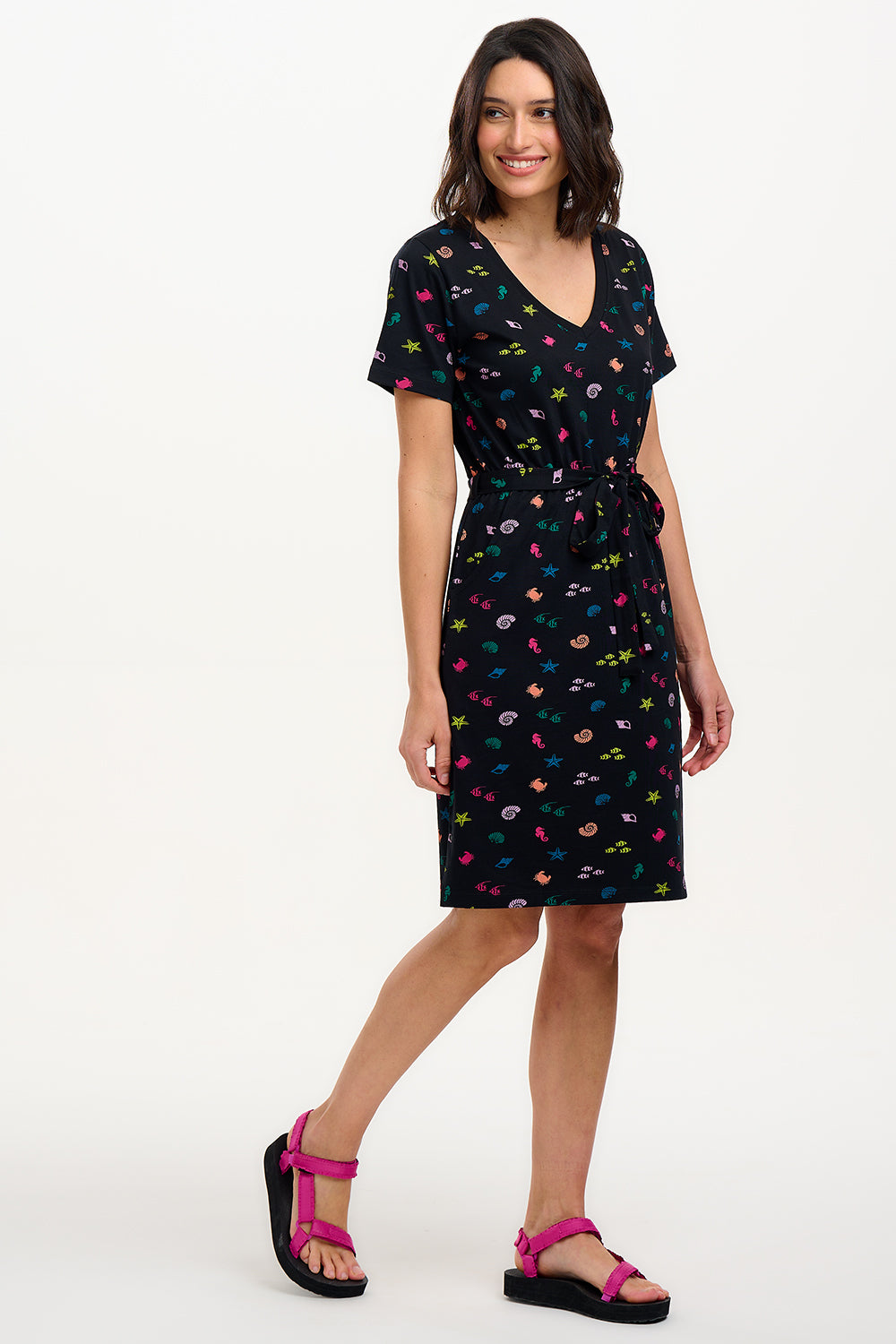 A model wearing a short-sleeved black mini dress with a V-neck, featuring a multicolored underwater theme print. The dress has a matching tie belt and pockets on the skirt.