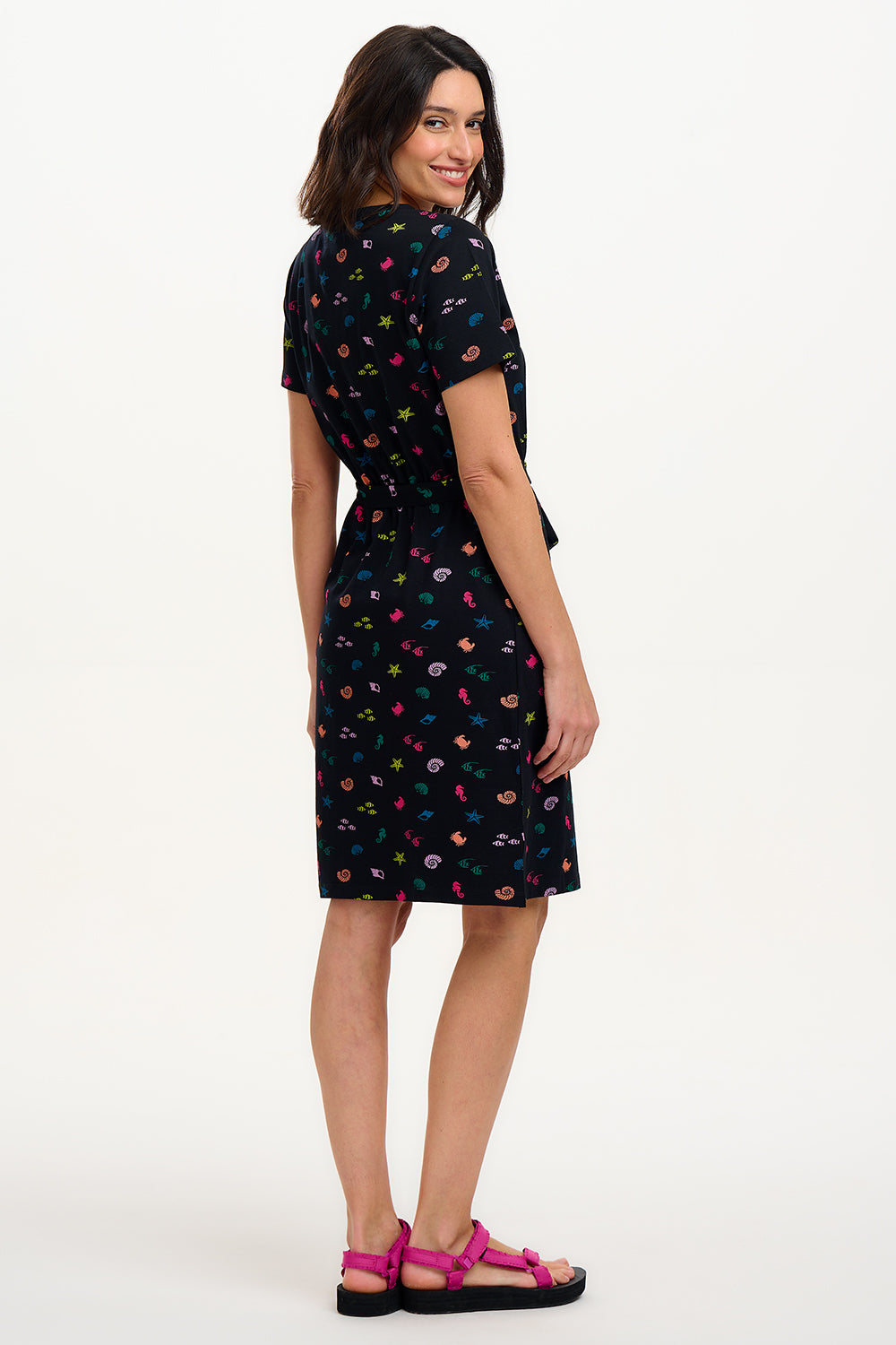 A model wearing a short-sleeved black mini dress with a V-neck, featuring a multicolored underwater theme print. The dress has a matching tie belt and pockets on the skirt.