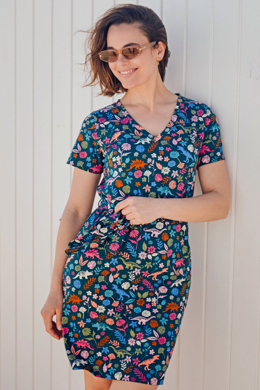 A woman modeling a teal and multicolored floral mini dress with a V-neck and short sleeves, standing against a white wooden background.