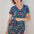 A woman modeling a teal and multicolored floral mini dress with a V-neck and short sleeves, standing against a white wooden background.