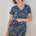 A woman modeling a teal and multicolored floral mini dress with a V-neck and short sleeves, standing against a white wooden background.