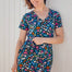 A woman modeling a teal and multicolored floral mini dress with a V-neck and short sleeves, standing against a white wooden background.