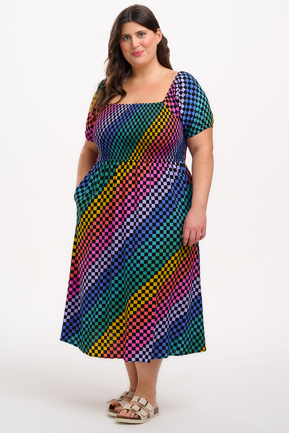 A model wearing a square neckline, short sleeves, high waist, and full midi skirt with pockets, featuring a black base with a multicolored rainbow checkerboard pattern.