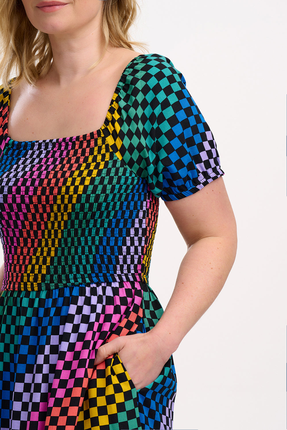 A model wearing a square neckline, short sleeves, high waist, and full midi skirt with pockets, featuring a black base with a multicolored rainbow checkerboard pattern.