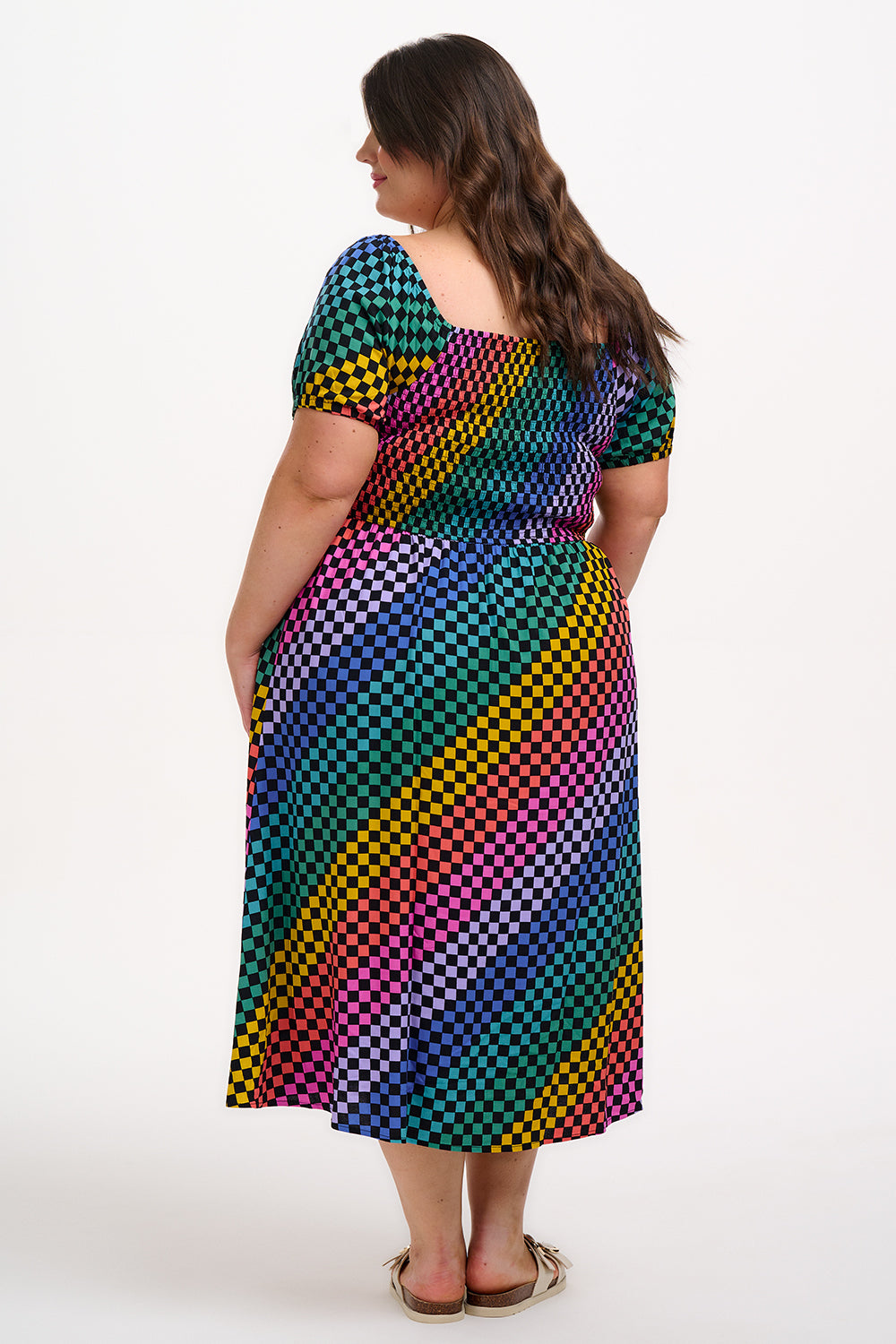 A model wearing a square neckline, short sleeves, high waist, and full midi skirt with pockets, featuring a black base with a multicolored rainbow checkerboard pattern.
