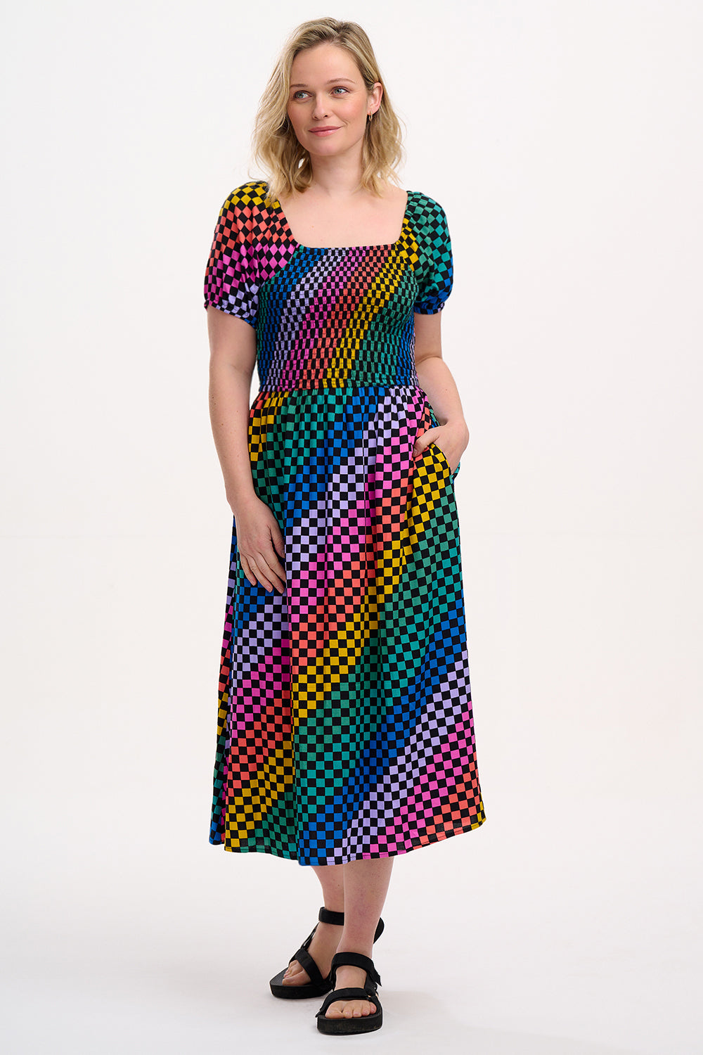 A model wearing a square neckline, short sleeves, high waist, and full midi skirt with pockets, featuring a black base with a multicolored rainbow checkerboard pattern.