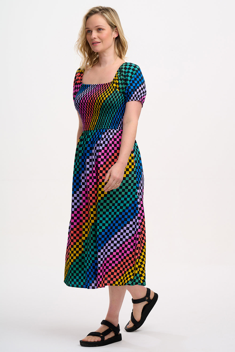 A model wearing a square neckline, short sleeves, high waist, and full midi skirt with pockets, featuring a black base with a multicolored rainbow checkerboard pattern.