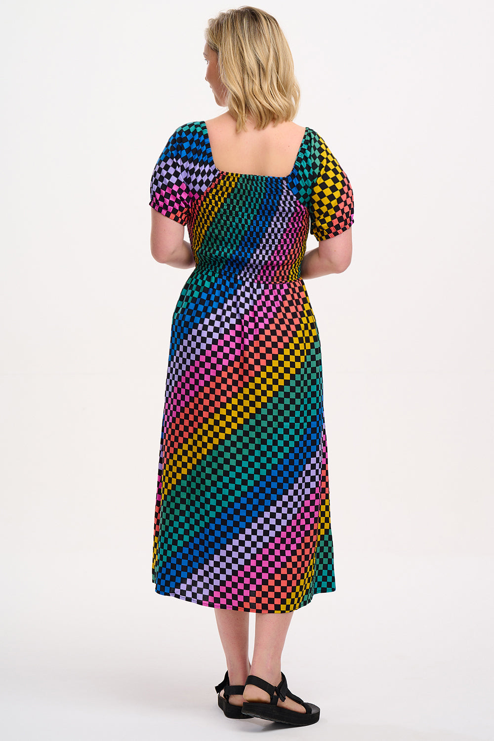 A model wearing a square neckline, short sleeves, high waist, and full midi skirt with pockets, featuring a black base with a multicolored rainbow checkerboard pattern.