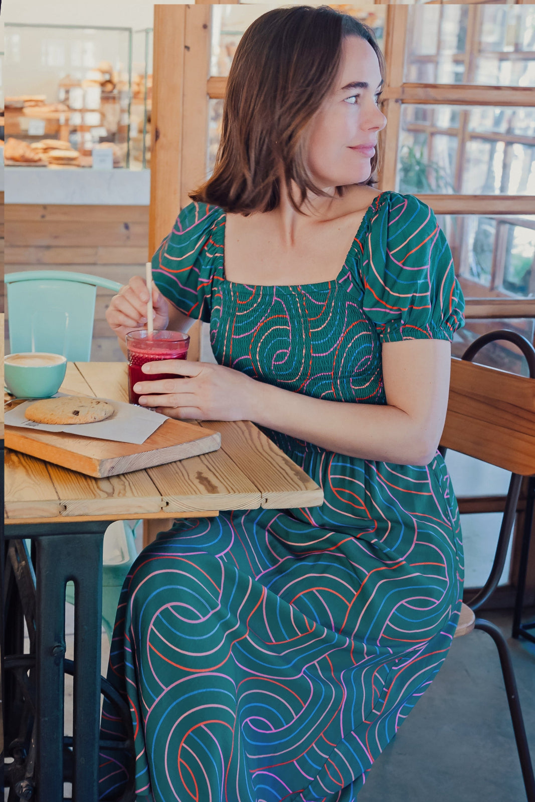 A woman sitting at a table wearing a square neckline, short sleeves, high waist, and midi length green dress with a geometric concentric circles pattern.