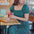 A woman sitting at a table wearing a square neckline, short sleeves, high waist, and midi length green dress with a geometric concentric circles pattern.