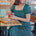 A woman sitting at a table wearing a square neckline, short sleeves, high waist, and midi length green dress with a geometric concentric circles pattern.