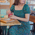 A woman sitting at a table wearing a square neckline, short sleeves, high waist, and midi length green dress with a geometric concentric circles pattern.