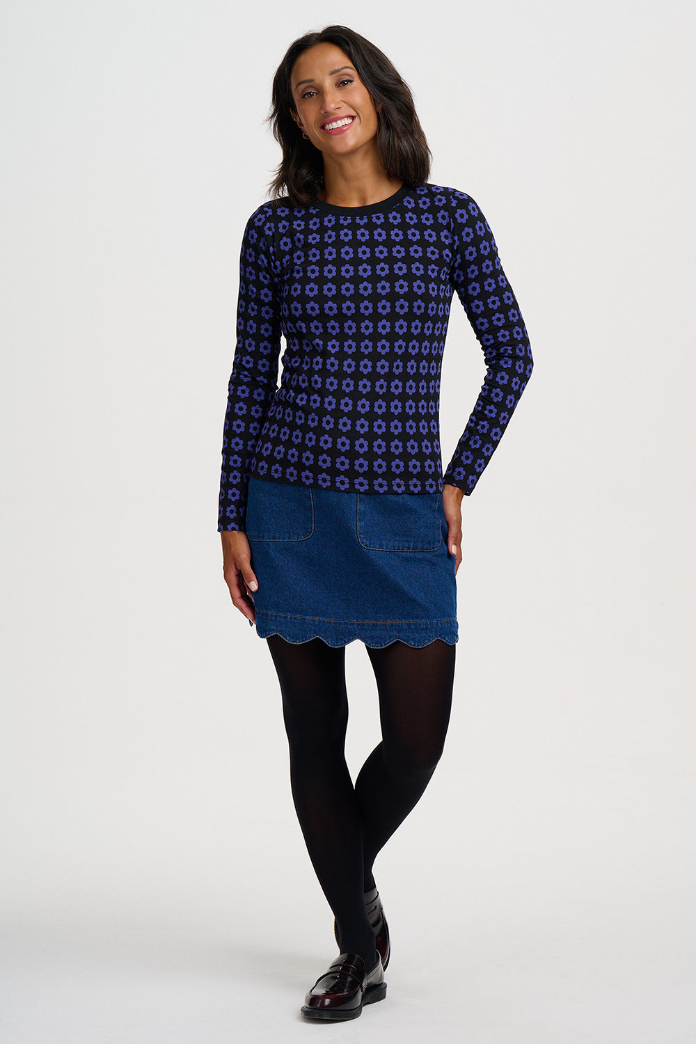 A woman modeling a navy blue long-sleeve top with a small geometric pattern, paired with a blue denim mini skirt, black tights, and black shoes, against a light gray studio background.