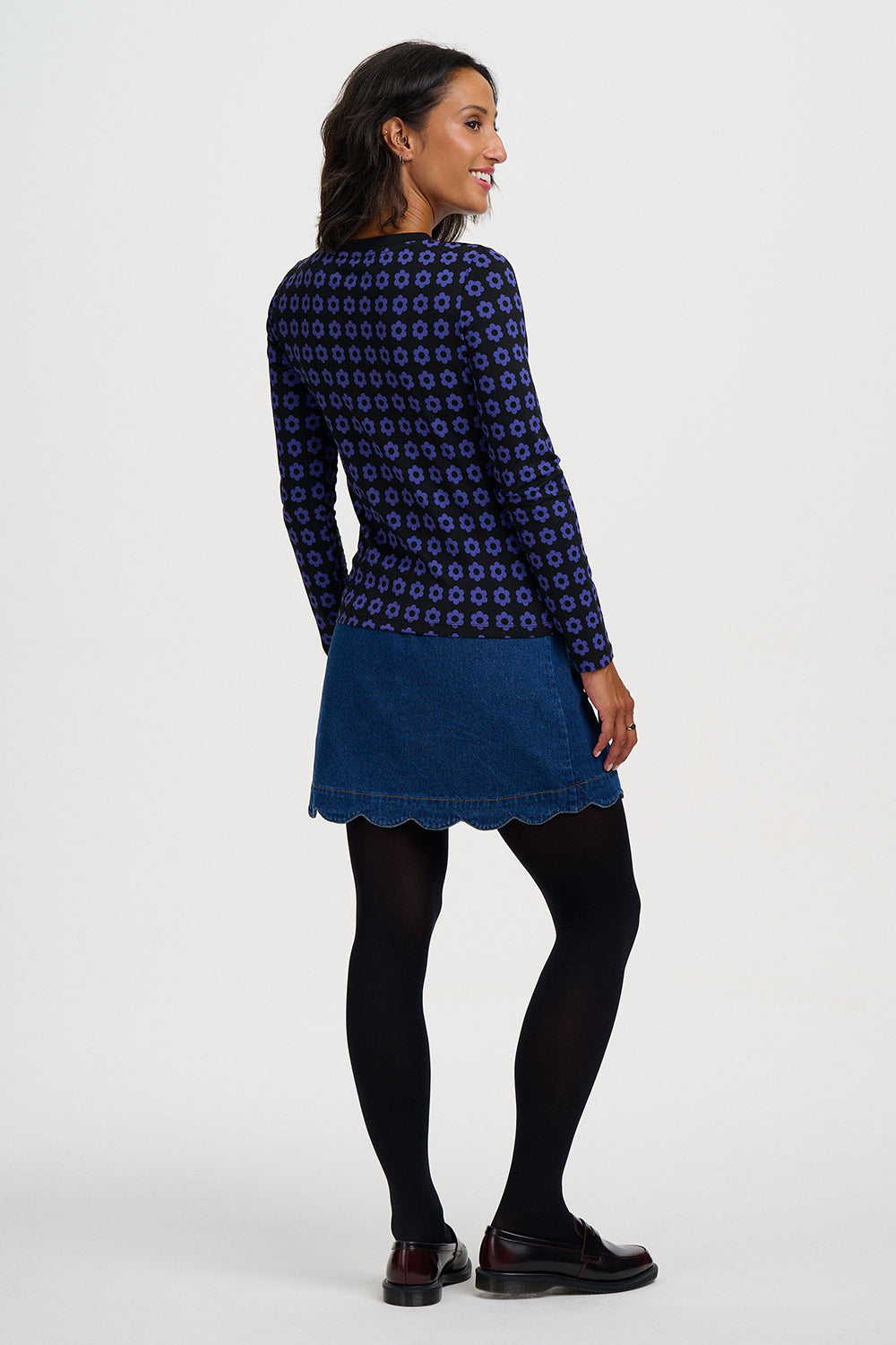 A woman is shown from the back wearing a navy blue long-sleeve top with a small geometric pattern, a blue denim mini skirt with a scalloped hem, black tights, and black shoes, against a plain light gray studio background.