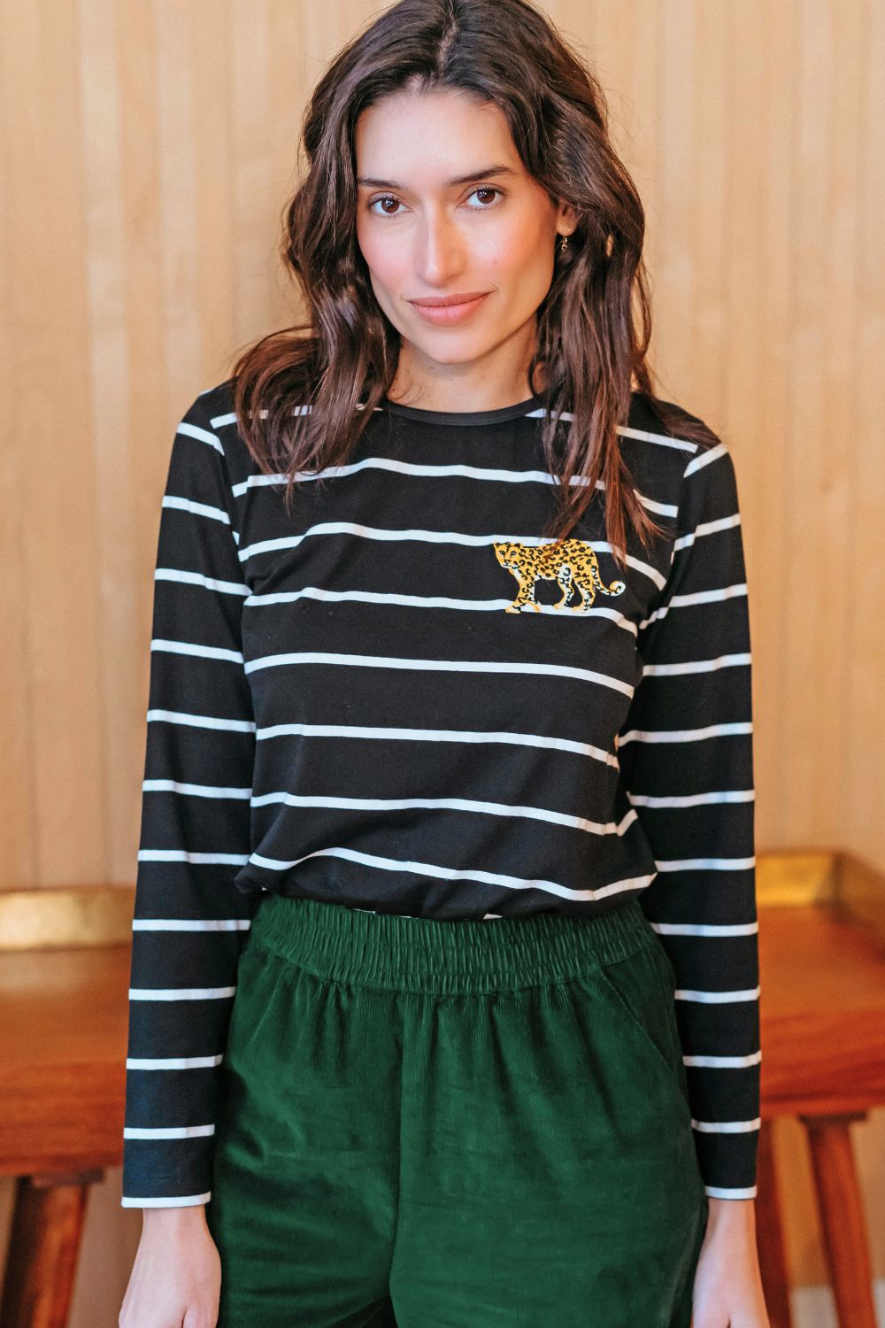 Sugarhill Brighton model wearing a black and white striped shirt with a gold emblem and green pants indoors.