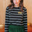 Sugarhill Brighton model wearing a black and white striped shirt with a gold emblem and green pants indoors.