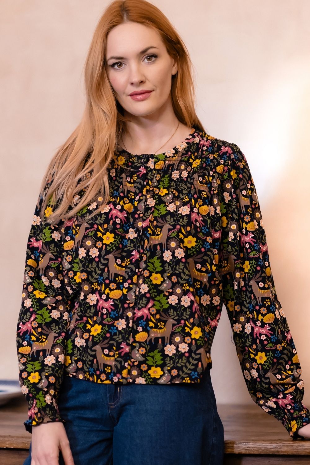 A woman with long red hair wearing a long-sleeve black blouse with a colorful floral and deer pattern, paired with dark blue jeans, standing against a warm beige background.