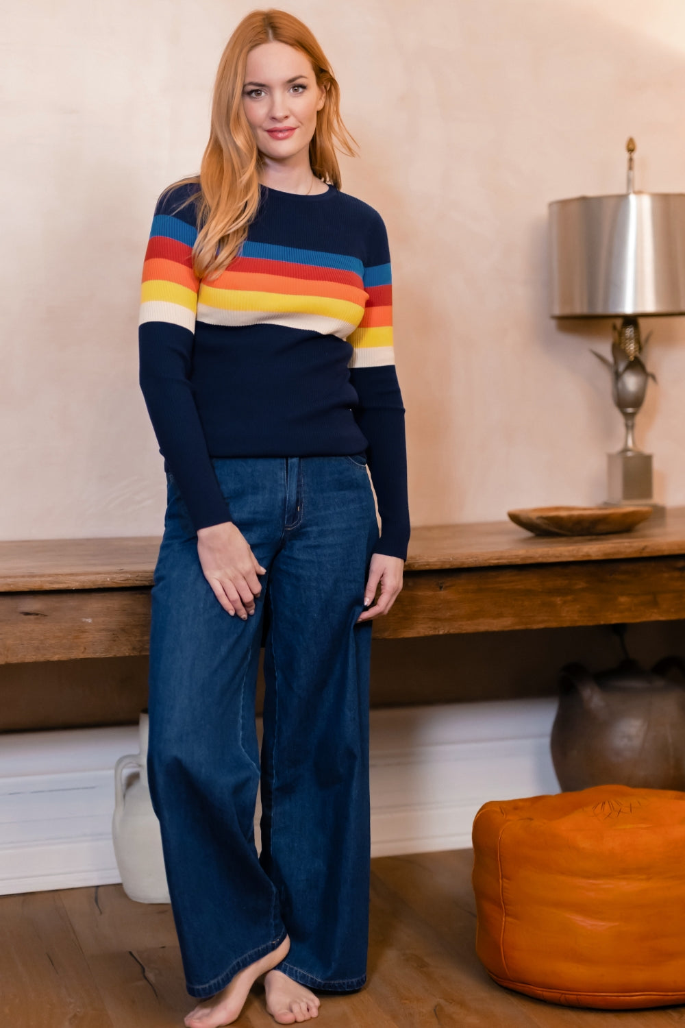 A woman stands barefoot in a room wearing a navy blue knit sweater with horizontal rainbow stripes across the chest and sleeves, paired with wide-leg blue jeans.