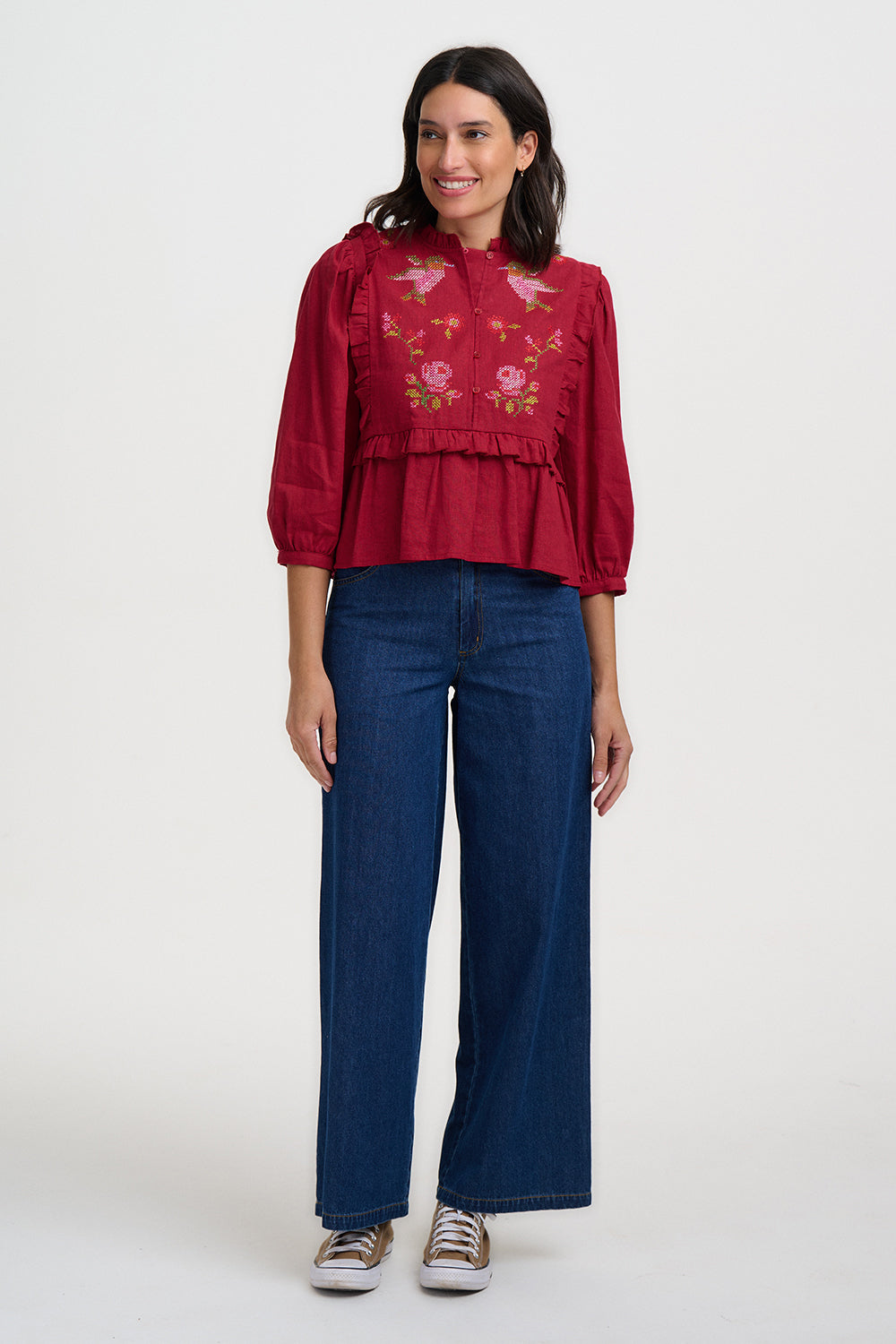 A woman modeling a red long-sleeve blouse with floral embroidery on the chest, paired with dark blue wide-leg jeans and brown sneakers, against a plain white studio background.