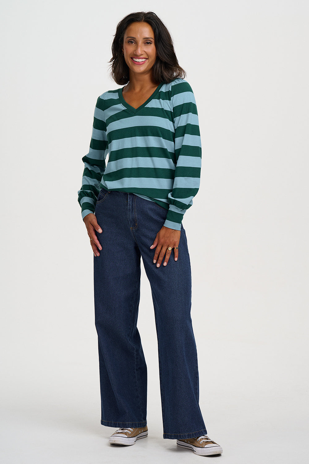 A woman modeling a green and light blue striped long-sleeve V-neck top with dark blue wide-leg jeans and brown sneakers, standing against a light gray studio background.