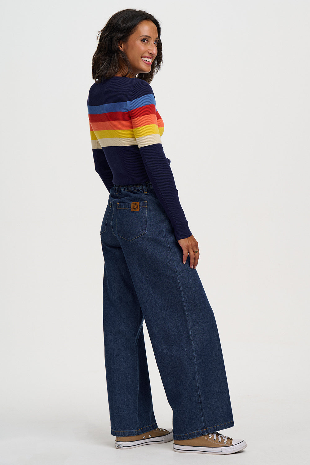 A woman modeling a navy blue knit sweater with horizontal rainbow stripes (red, orange, yellow, cream, blue) and dark blue wide-leg denim jeans, standing against a light gray studio background.