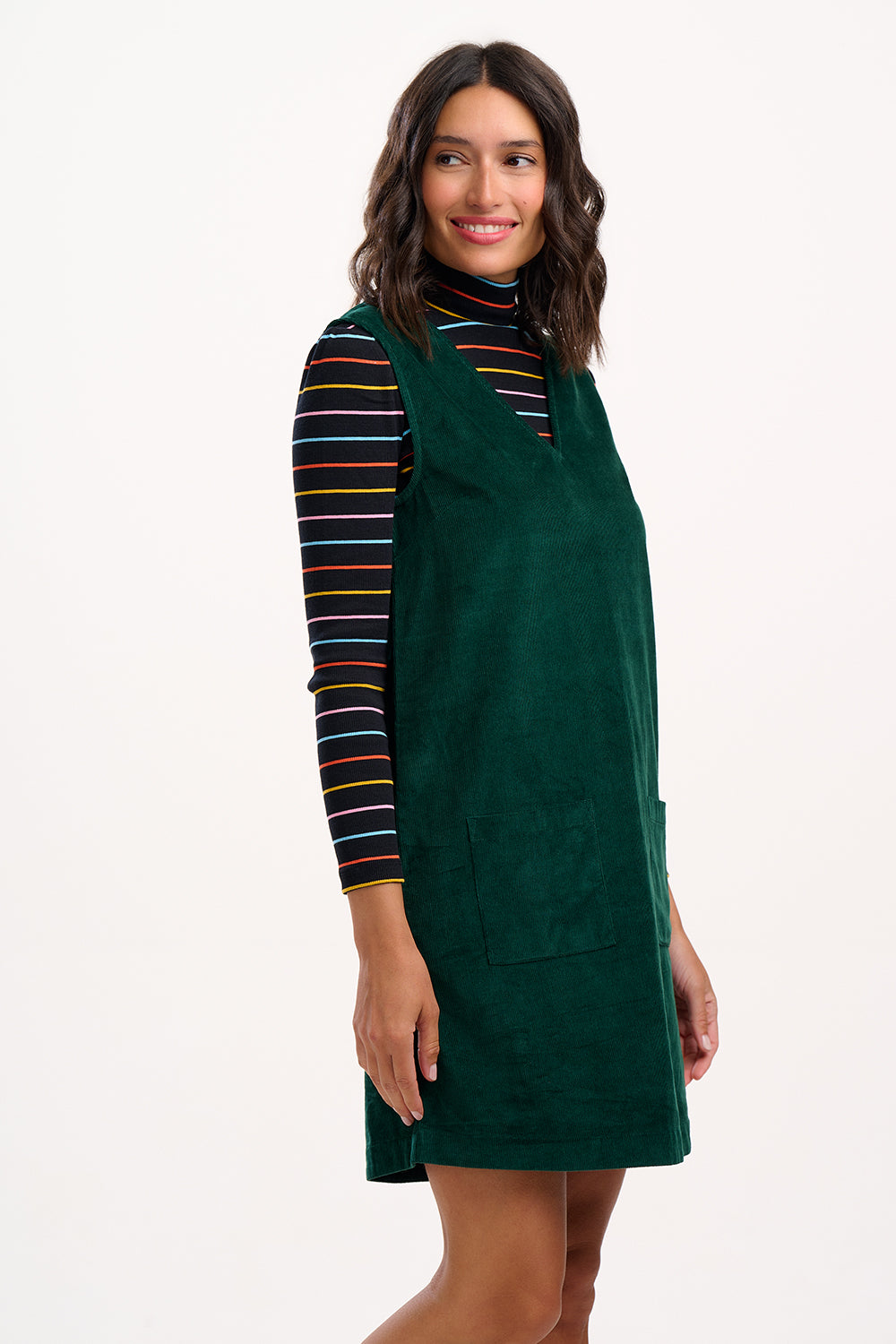 Woman wearing a green pinafore dress over a striped shirt on a white background