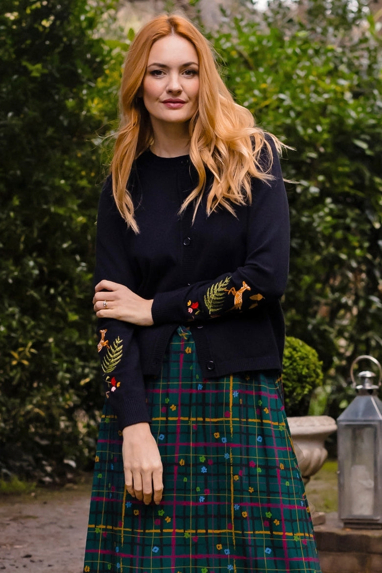 A woman stands outdoors wearing a dark navy cardigan with colorful embroidered leaf details on the sleeves and a green tartan plaid midi skirt with small multicolored embroidered dots. The background features green hedges, a stone lantern, and a gravel path.