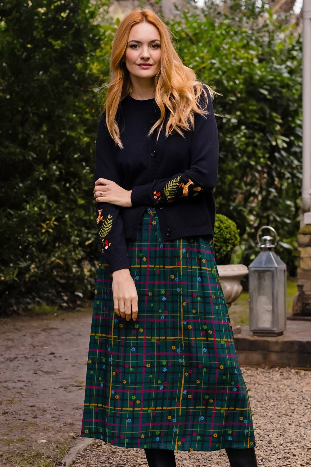A woman stands outdoors wearing a dark navy cardigan with colorful embroidered leaf details on the sleeves and a green tartan plaid midi skirt with small multicolored embroidered dots. The background features green hedges, a stone lantern, and a gravel path.