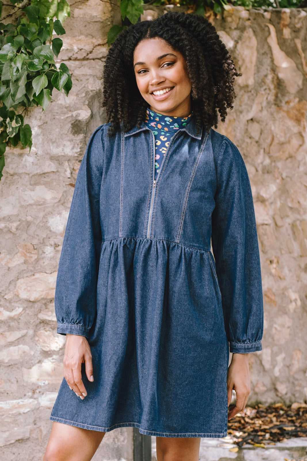 A woman wearing a short blue denim smock dress with a classic shirt collar and long cuffed sleeves.