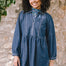 A woman wearing a short blue denim smock dress with a classic shirt collar and long cuffed sleeves.