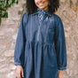 A woman wearing a short blue denim smock dress with a classic shirt collar and long cuffed sleeves.
