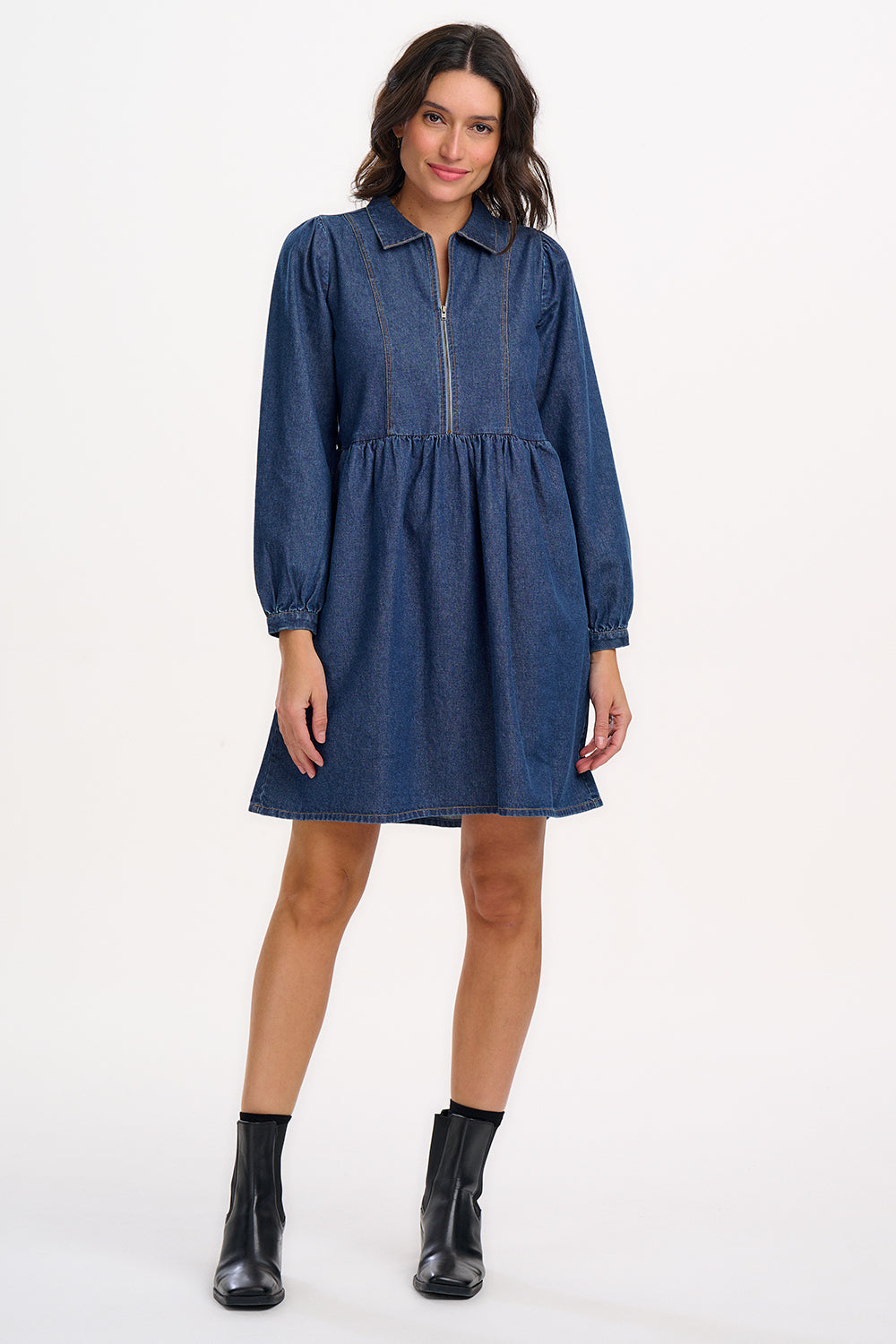 A woman wearing a short blue denim smock dress with a classic shirt collar and long cuffed sleeves.