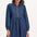 A woman wearing a short blue denim smock dress with a classic shirt collar and long cuffed sleeves.