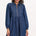 A woman wearing a short blue denim smock dress with a classic shirt collar and long cuffed sleeves.