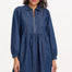 A woman wearing a short blue denim smock dress with a classic shirt collar and long cuffed sleeves.