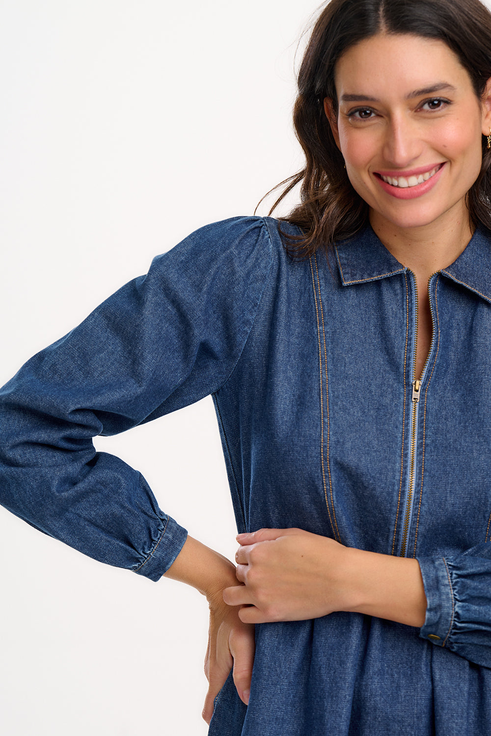 A woman wearing a short blue denim smock dress with a classic shirt collar and long cuffed sleeves.