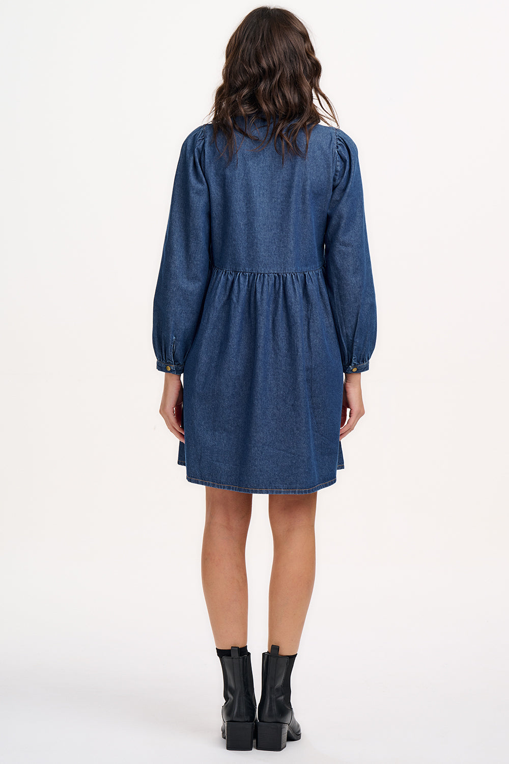 A woman wearing a short blue denim smock dress with a classic shirt collar and long cuffed sleeves.