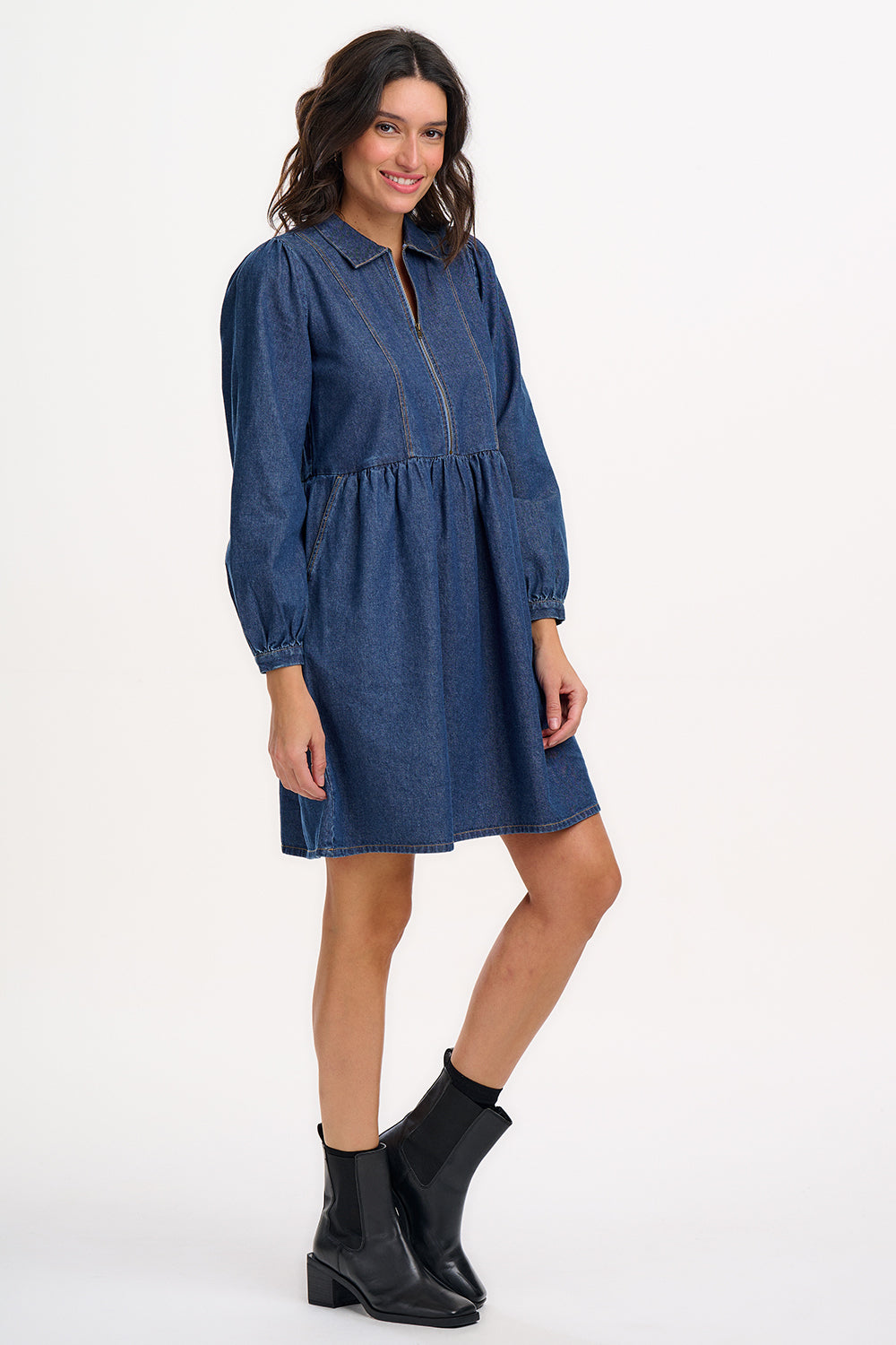 A woman wearing a short blue denim smock dress with a classic shirt collar and long cuffed sleeves.