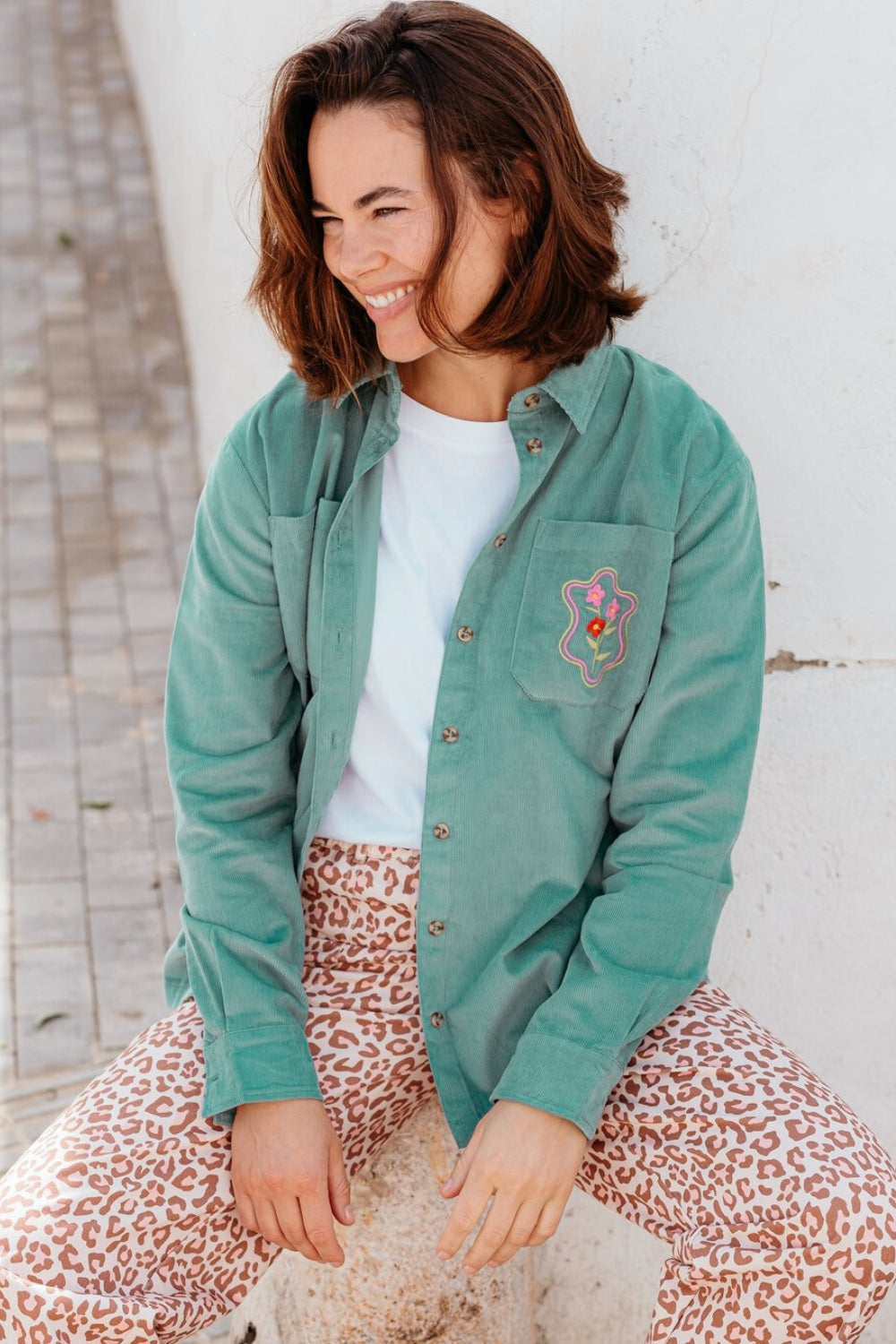Woman wearing a green jacket with a floral design sitting against a white wall.