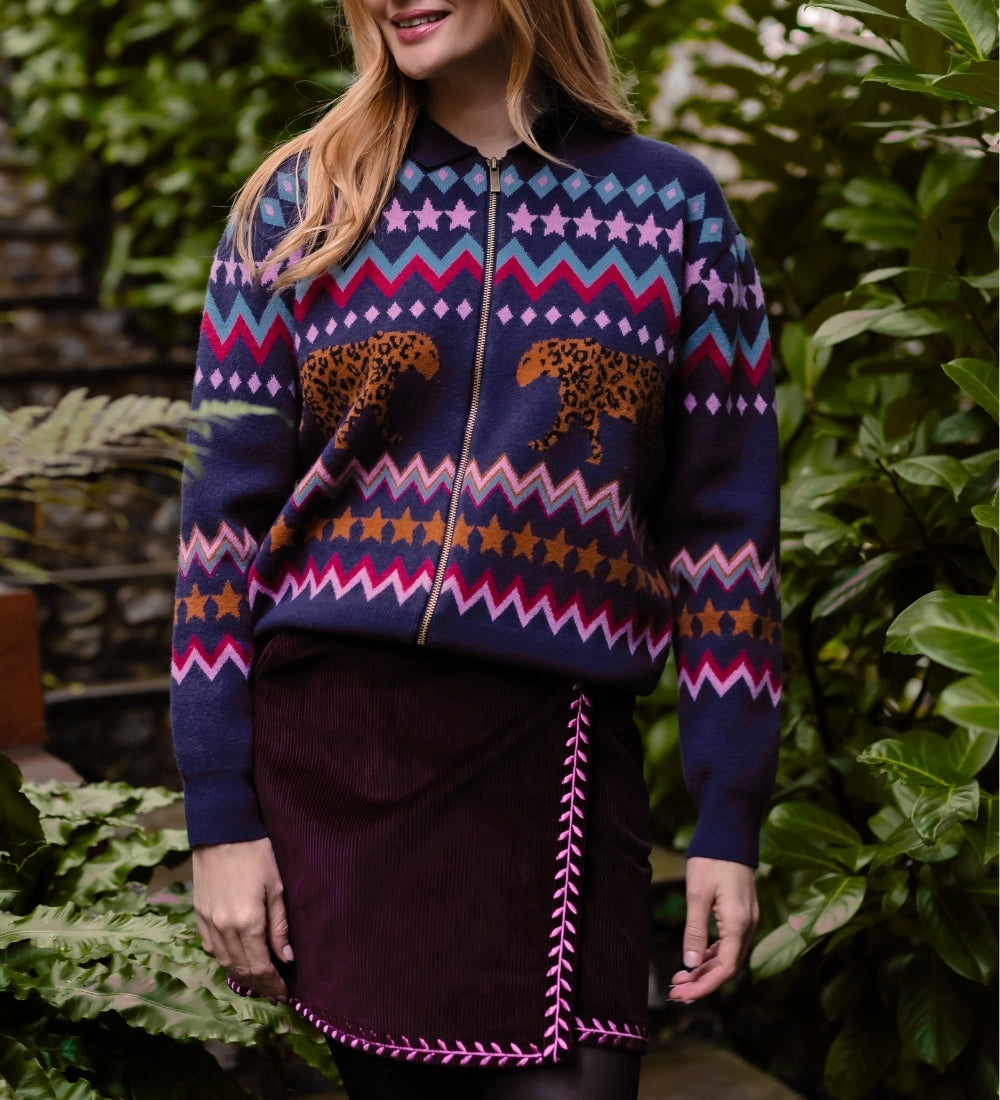 A woman wearing a navy blue zip-up sweater with a colorful Fair Isle pattern of stars, zigzags, and cheetahs, paired with a dark purple skirt, standing in front of a lush green garden background.