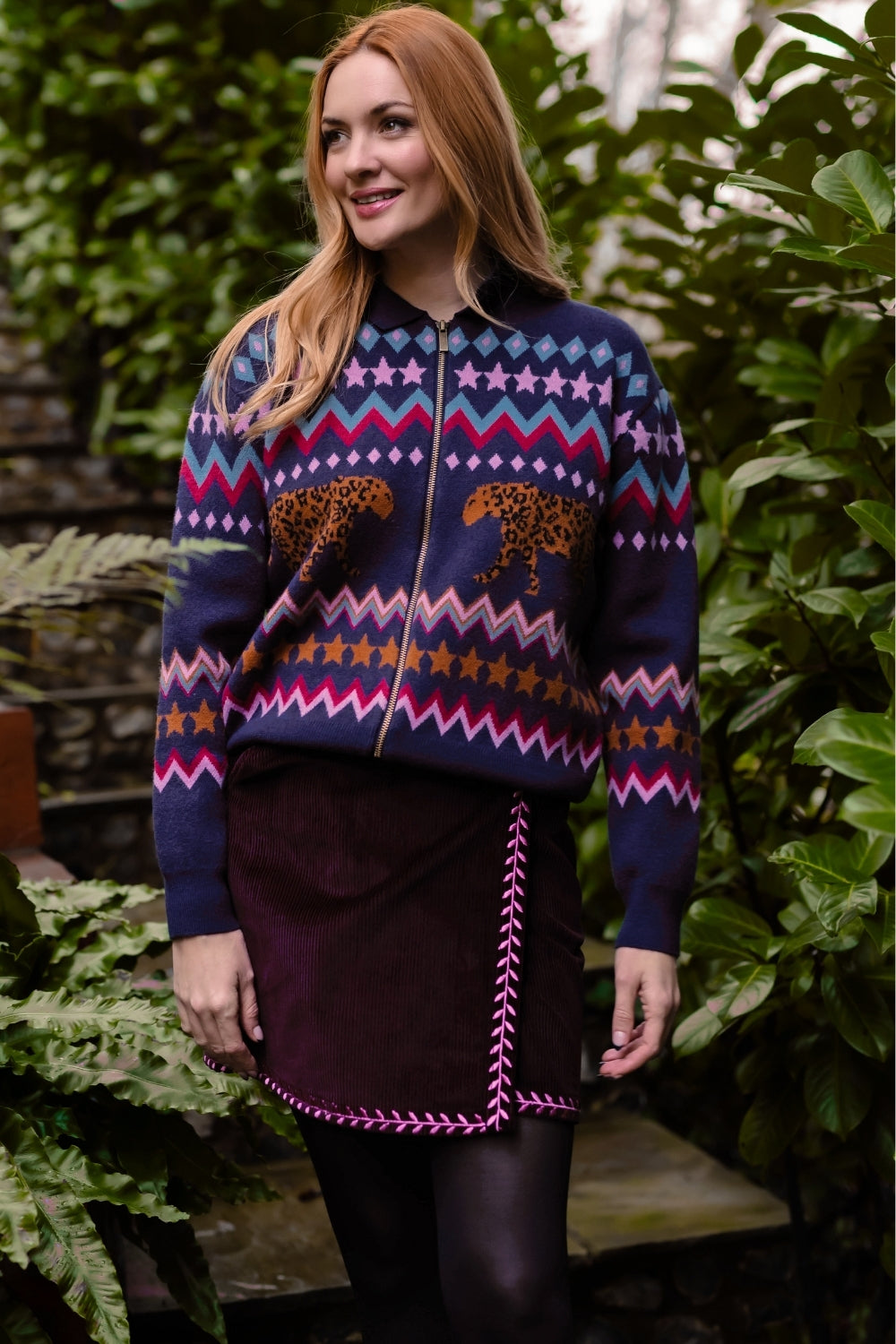 A woman wearing a navy blue zip-up sweater with a colorful Fair Isle pattern of stars, zigzags, and cheetahs, paired with a dark purple skirt, standing in front of a lush green garden background.
