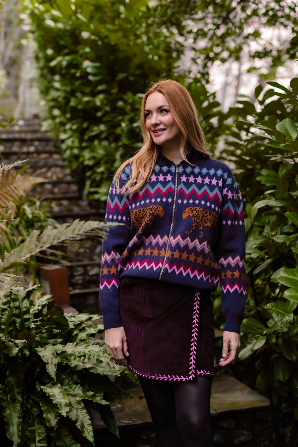 A woman with red hair stands outdoors in a lush green garden setting, wearing a navy blue Fair Isle patterned zip-up sweater with leopard print details and a dark purple skirt.