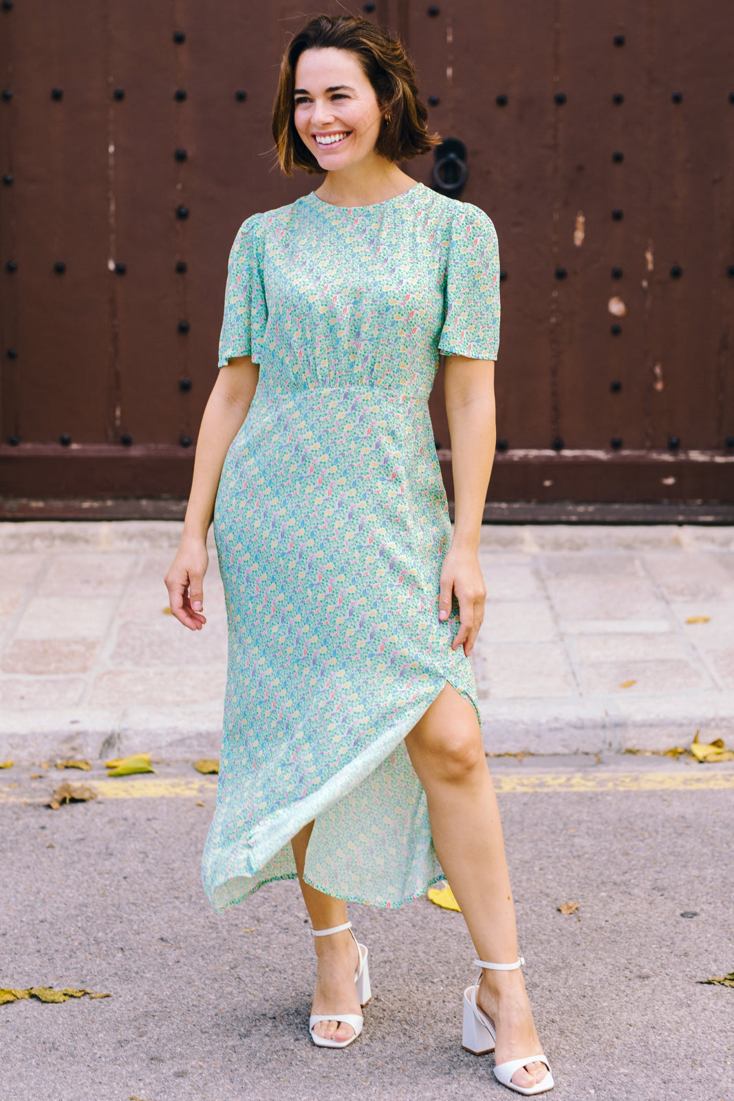 A woman wearing an off-white midi tea dress with a Garden Waves print, featuring short fluted sleeves and a slit skirt, standing outdoors.