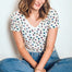 A woman sitting cross-legged and wearing a beige V-neck T-shirt with a multicolor polka dot pattern, paired with blue jeans.