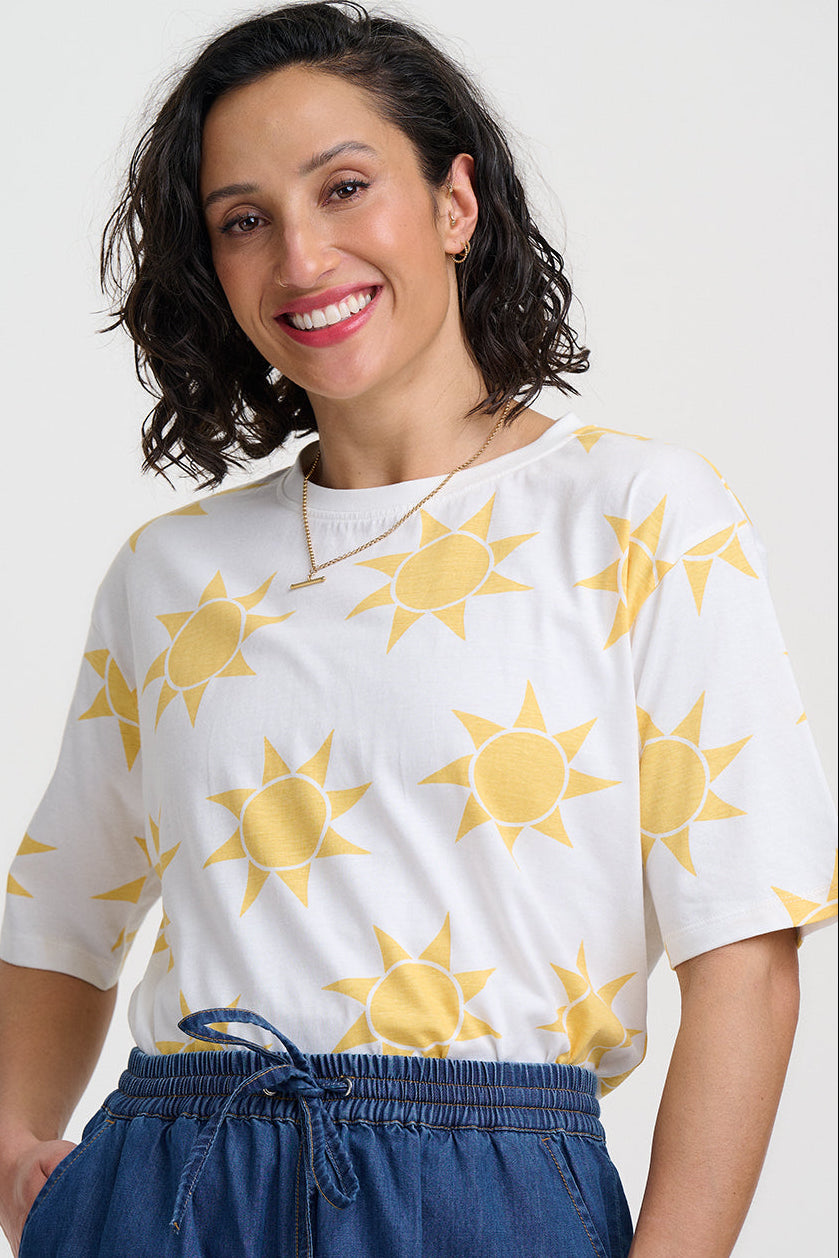 Woman wearing a white shirt with yellow sun patterns and blue pants on a light gray background