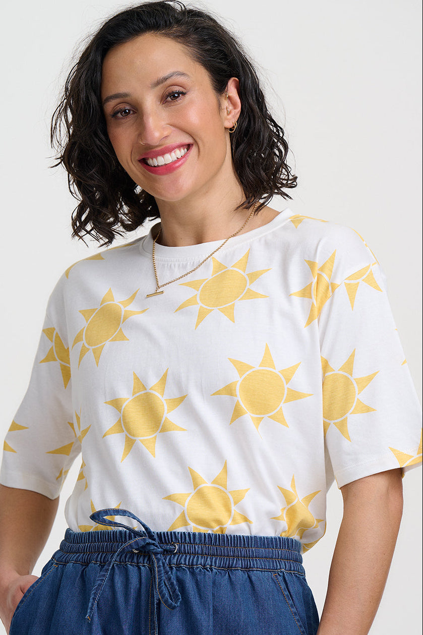 Woman wearing a white shirt with yellow sun patterns and blue pants on a light gray background