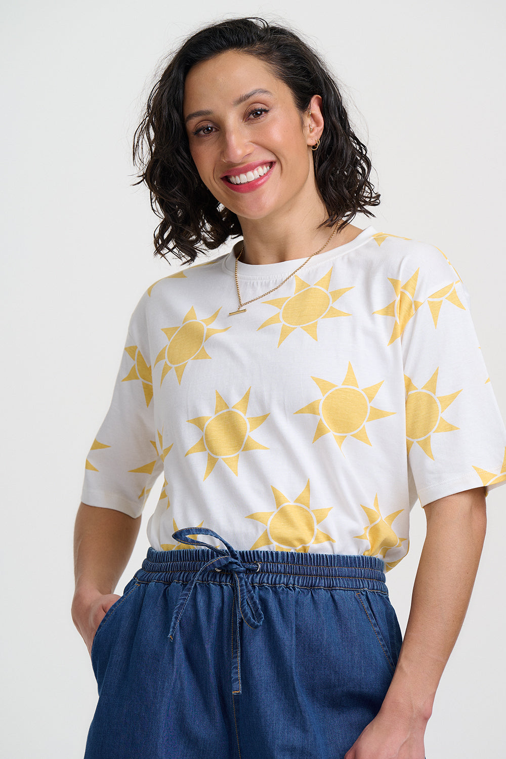 Woman wearing a white shirt with yellow sun patterns and blue pants on a light gray background