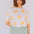 Woman wearing a white shirt with yellow sun patterns and light green pants against a white wall.